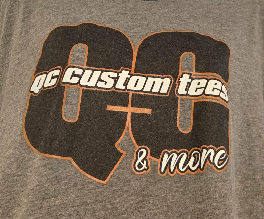 Close-up of QC Custom Tees logo on gray T-shirt fabric for custom apparel.