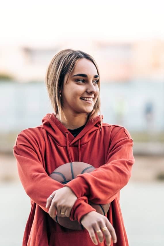 Woman in red hoodie holding basketball outdoors, smiling.