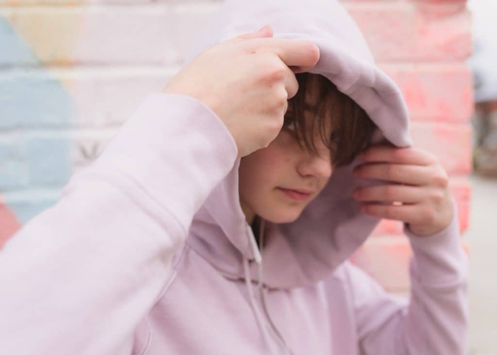 Person wearing a light purple hoodie adjusting the hood in front of a colorful wall.